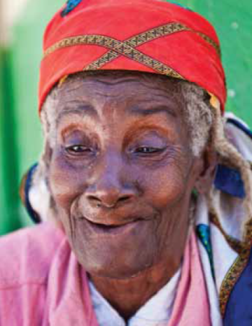 Elderly individual wearing a red headscarf and pink attire, smiling warmly.