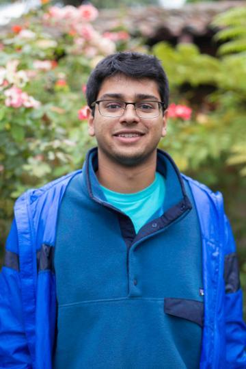 A person wearing a blue jacket standing in front of greenery.