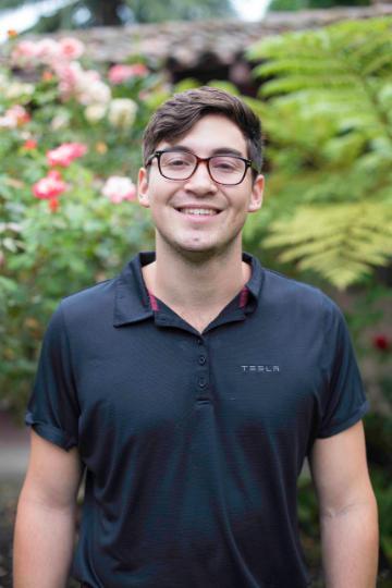 A person standing in a garden, smiling, wearing glasses and a dark shirt.