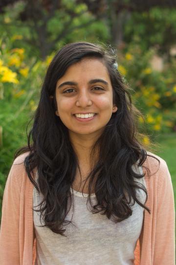 A person smiling outdoors with trees and flowers in the background.
