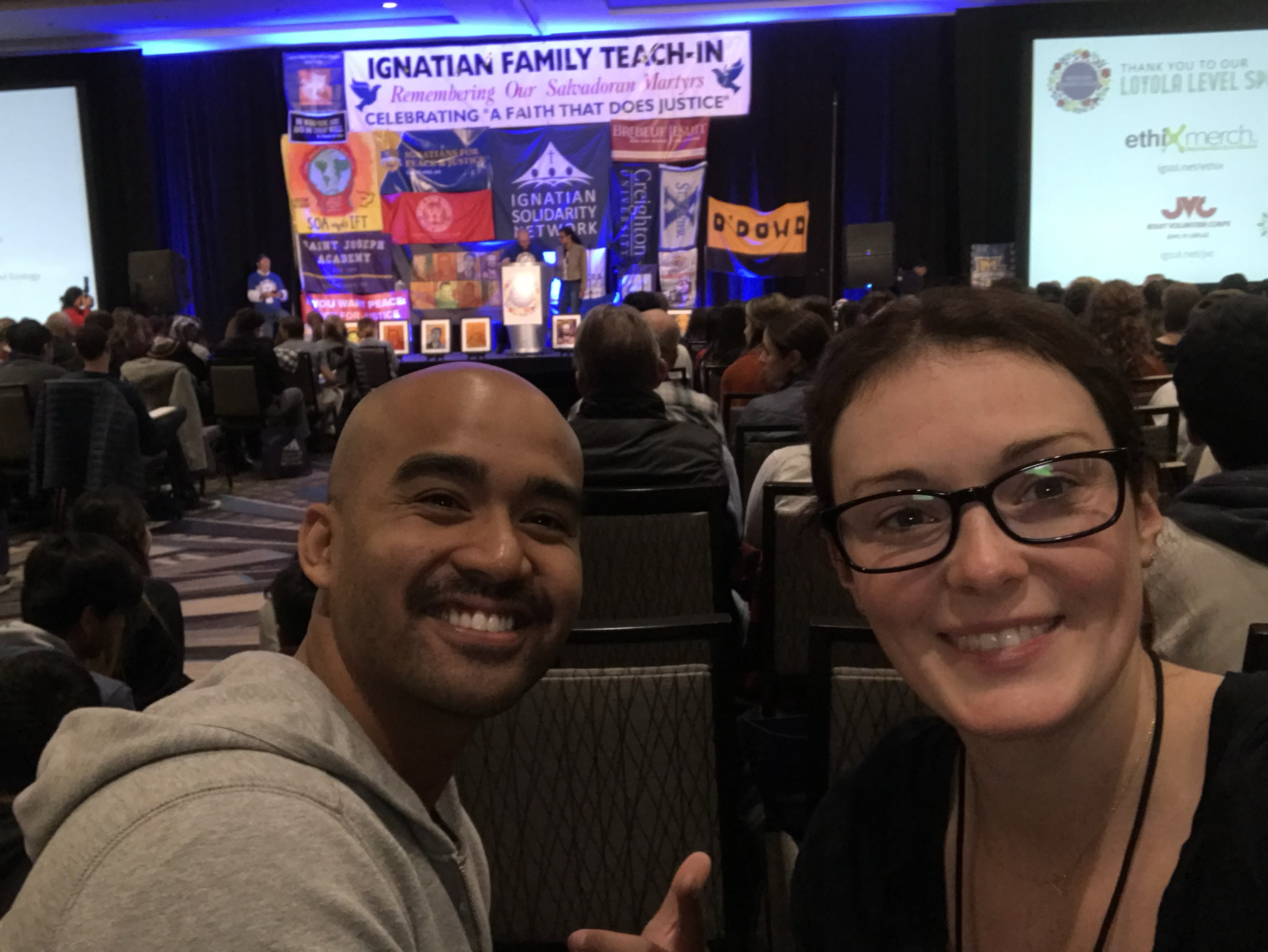 A man and woman smile in a conference hall with a lit stage.