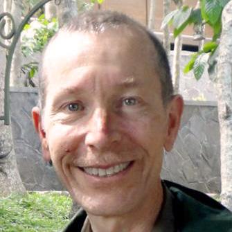 Man smiling outdoors with greenery and structures in the background.