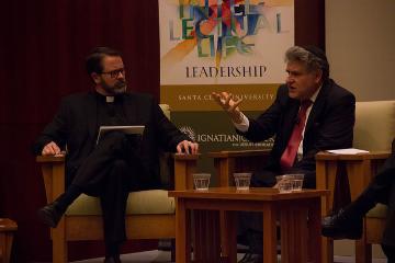 Two men in conversation seated on stage, colorful poster behind them.