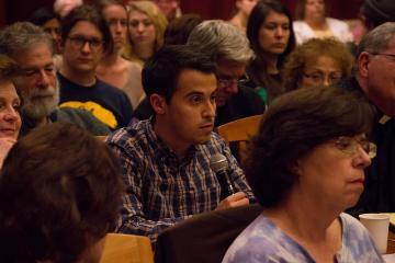 People seated and attentively listening in a crowded indoor event or gathering.