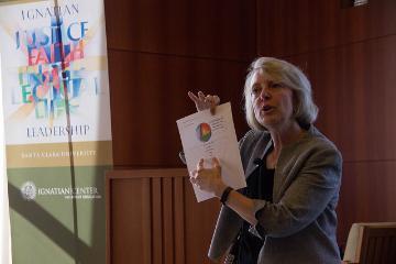 A person holding a booklet, speaking in a room with a colorful mural.