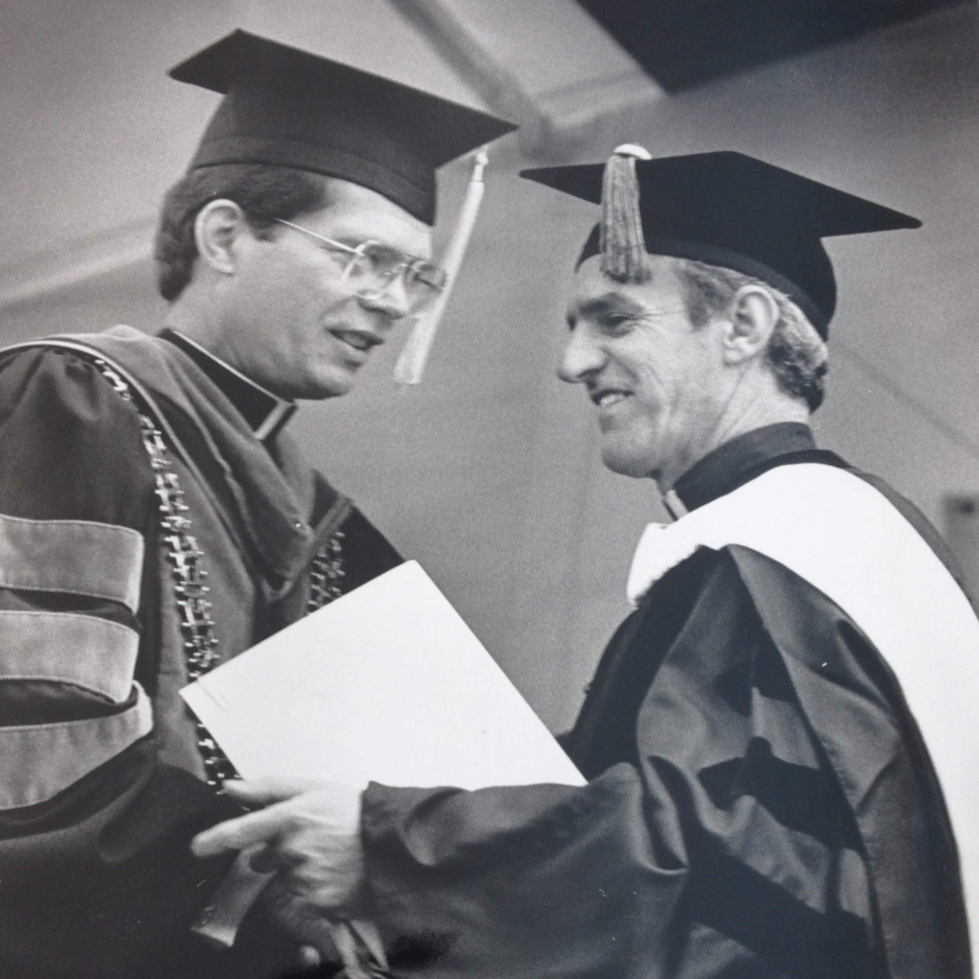 Two graduates in caps and gowns shake hands at commencement ceremony, 1982.