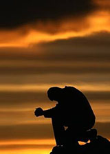 Silhouette of a person kneeling in prayer at sunset.