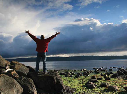 A person standing with arms raised near a lake under a cloudy sky.