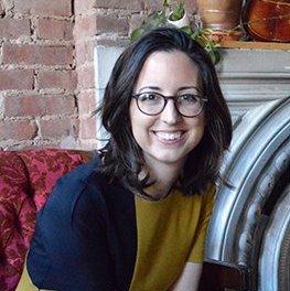 A person wearing glasses and smiling, seated indoors.