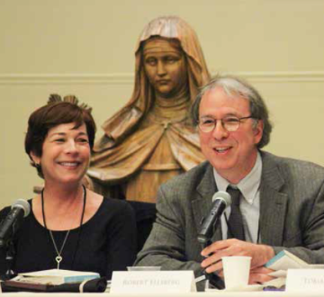Two people speaking at a panel with a statue in the background.