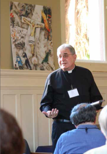 A man in clerical clothing giving a presentation, with people listening in the foreground.