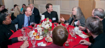 A group of people conversing around a table with a red tablecloth.