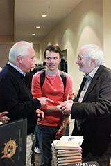 Three people are conversing; one is shaking hands in a hallway.