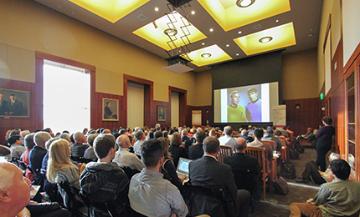 People seated at a conference or seminar with a speaker presenting on a screen.