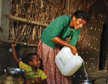 A woman pours water from a large container by a hut.