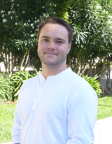 A person in a white shirt standing outdoors with greenery in the background.