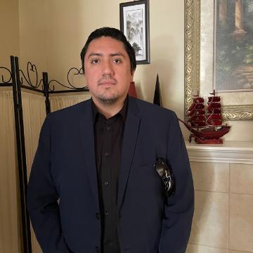 A person standing indoors wearing a suit, near a mantel with decorative items.