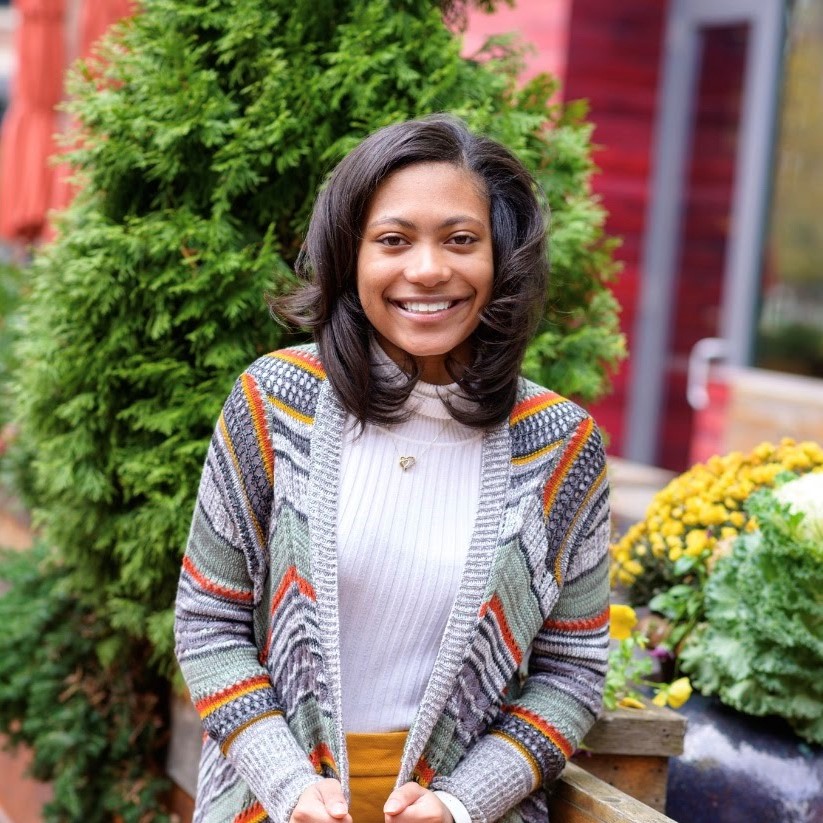 A woman smiling and holding a drink outdoors near plants. 