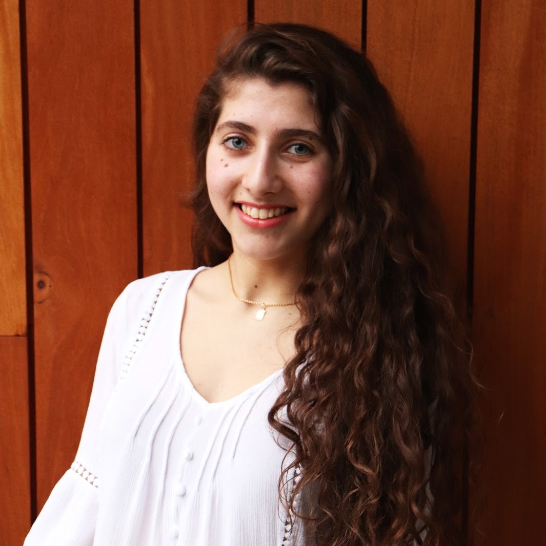 A person in a white blouse with long curly hair against a wooden wall. 