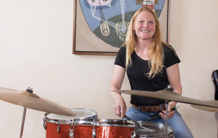 A woman holding drumsticks seated at a drum set with a jazz poster behind.
