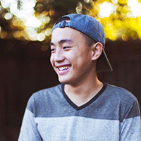A person smiling, wearing a gray shirt and hat, outdoors.