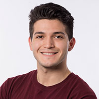 A person smiling, wearing a maroon shirt, with a plain background.