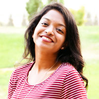 A woman smiling outdoors with blurred greenery in the background.