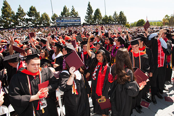 A large crowd of graduates in caps and gowns at an outdoor commencement ceremony.