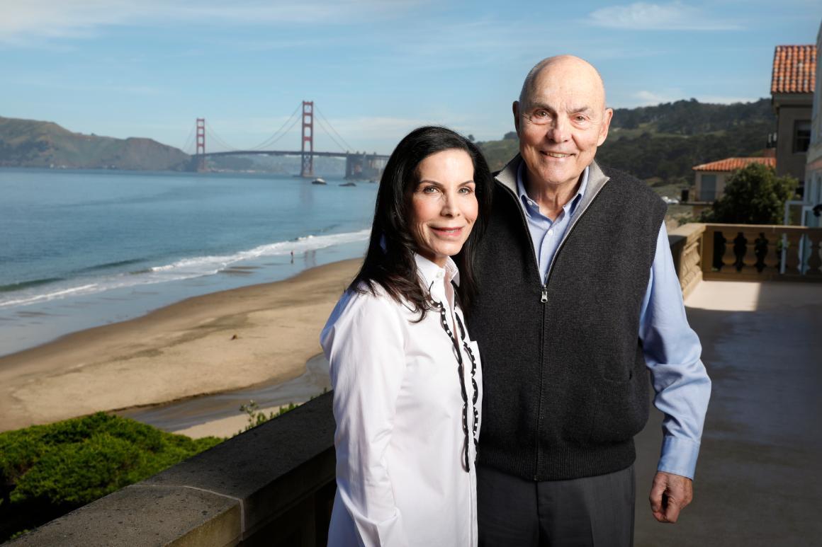 Two people standing in front of a waterfront bridge scene.