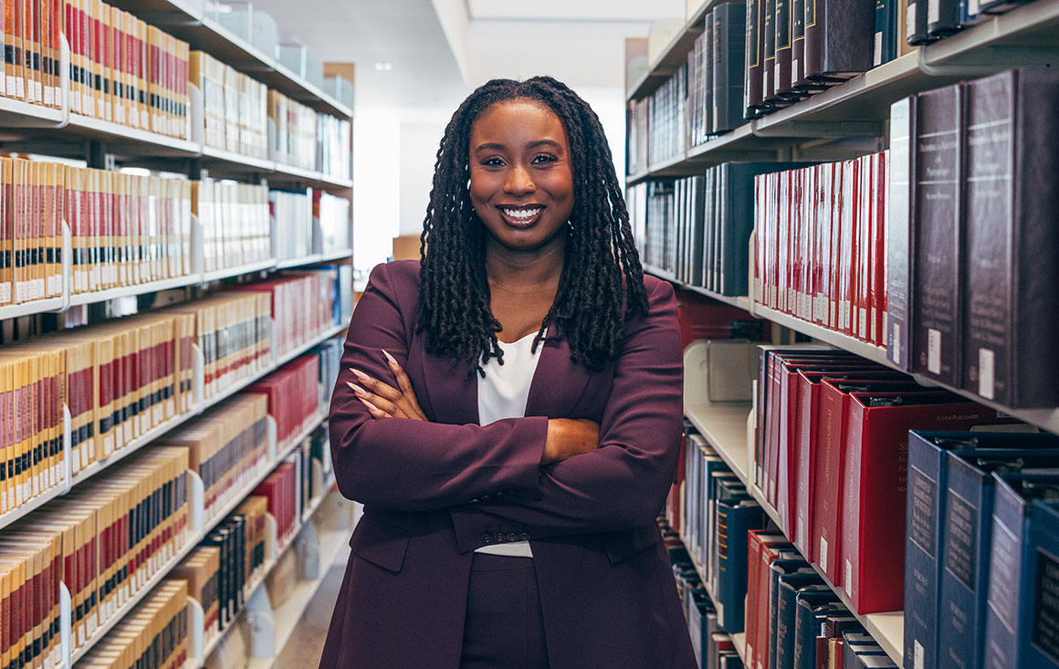 Ashanti Wallace smiles, stands with arms folded between stacks of books in the law library.