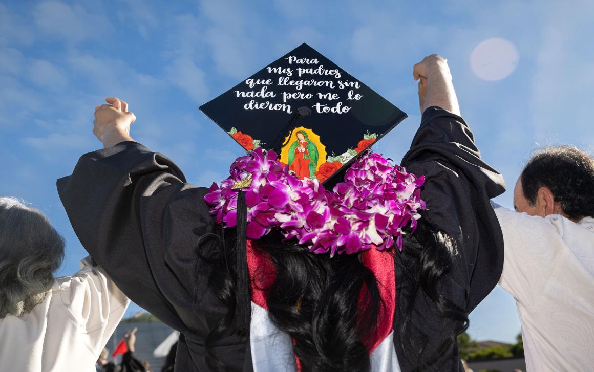 Graduate wearing a decorated cap with Spanish text and an image of the Virgin of Guadalupe, raising joined hands with parents at commencement.