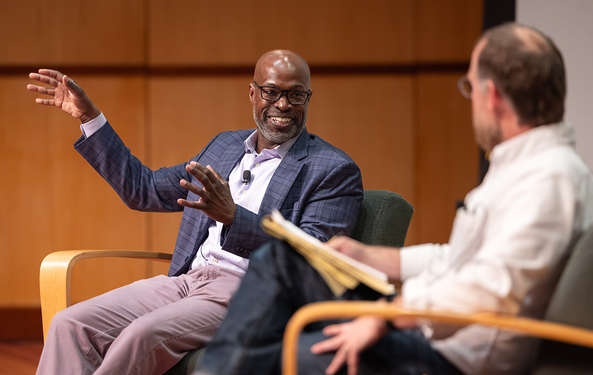 Kafui Dzirasa seated on stage in blue suit coat with his hand raised, speaking with blurred Santa Clara faculty member in foreground.