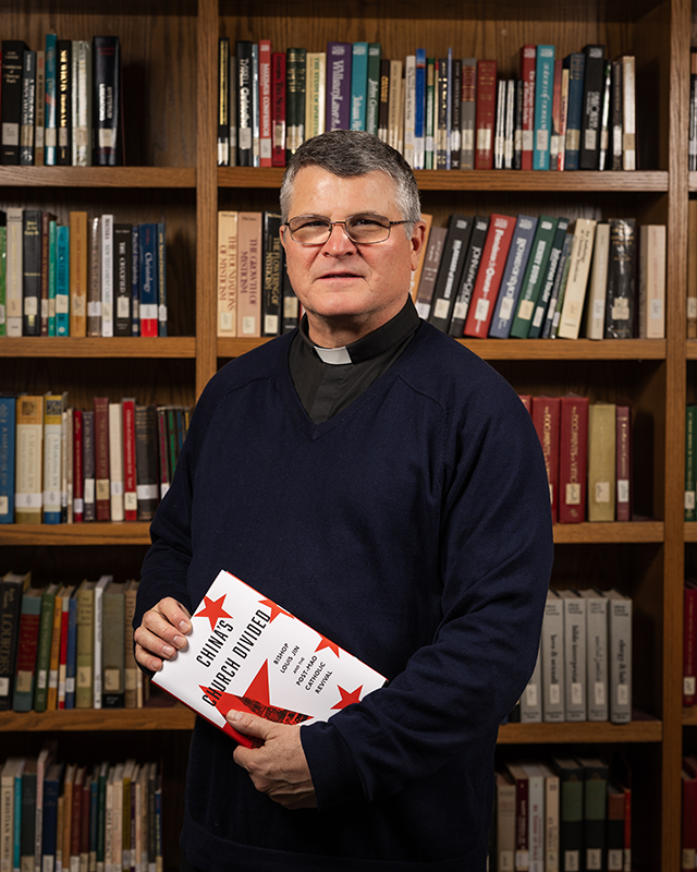 Paul Mariani holding book in front of library shelves in Jesuit Residences