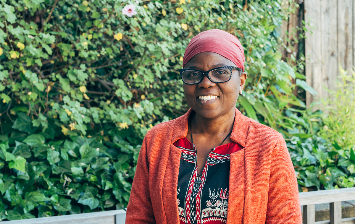 Woman wearing red wrap, black glasses, and orange cardigan standing in front of green bushes