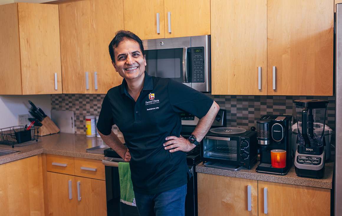 A Persian man stands with his hands playfully on his hips and a beaming smile in a kitchen, while brewing coffee.