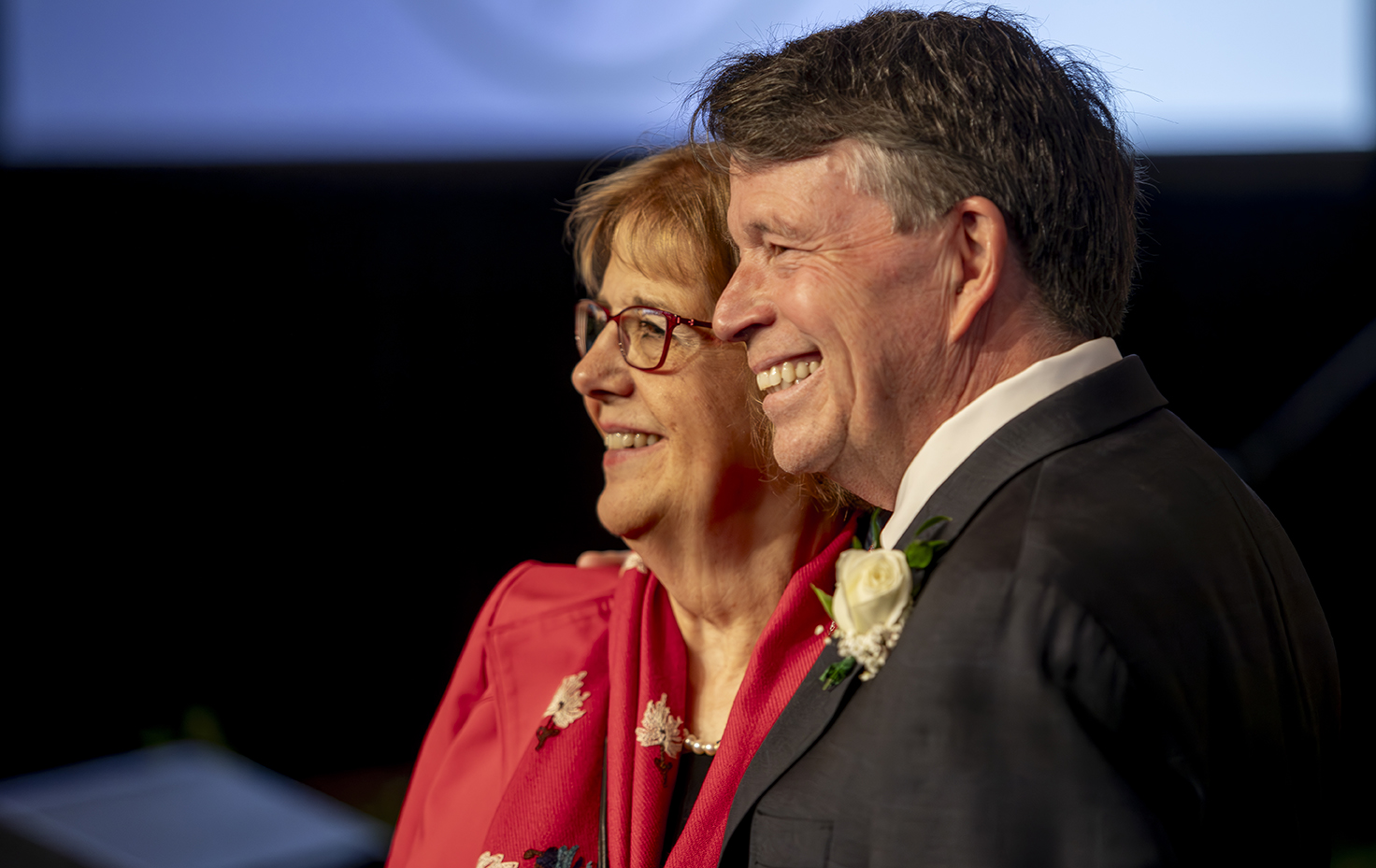 Silhouette of Jim Lyons on stage standing with a woman in a pink blazer.