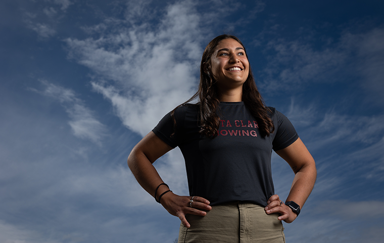 A student stands in a power pose with a cloudy blue sky behind them.