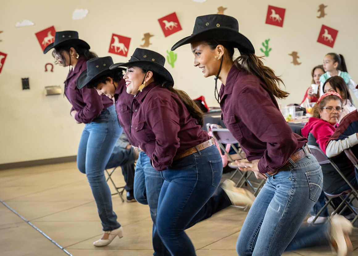 Four dancers in boots, denim jeans, and cowboy hats kick their feet while dancing.