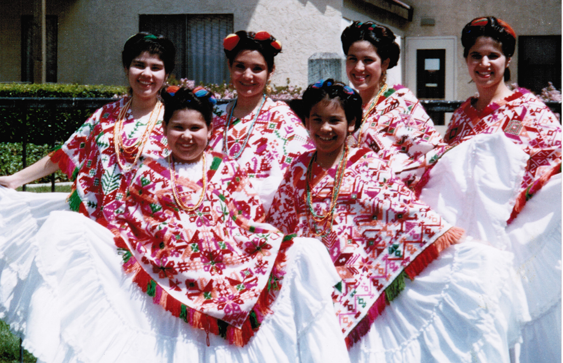 An older photograph of a group of young women in white, traditional Mexican dresses with decorative red ponchos.