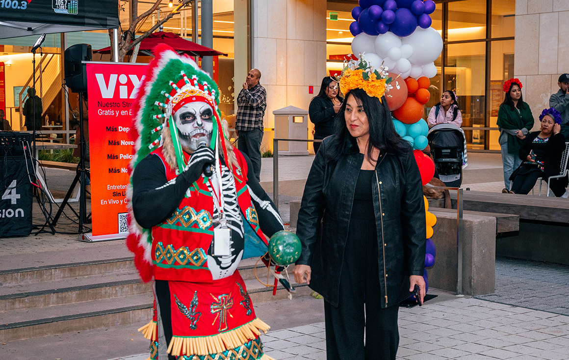 A man with skeleton face paint, a feathered headdress, and indigenous regalia holds a microphone while standing next to a Latina woman at a celebration.