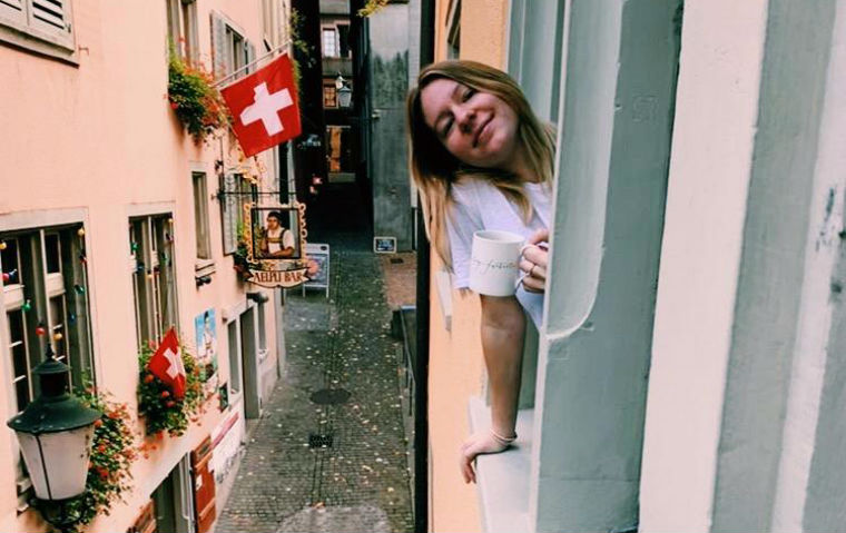 Study Abroad Student Sami Springman '21 leaning out a window over an alley with Swiss flags in background