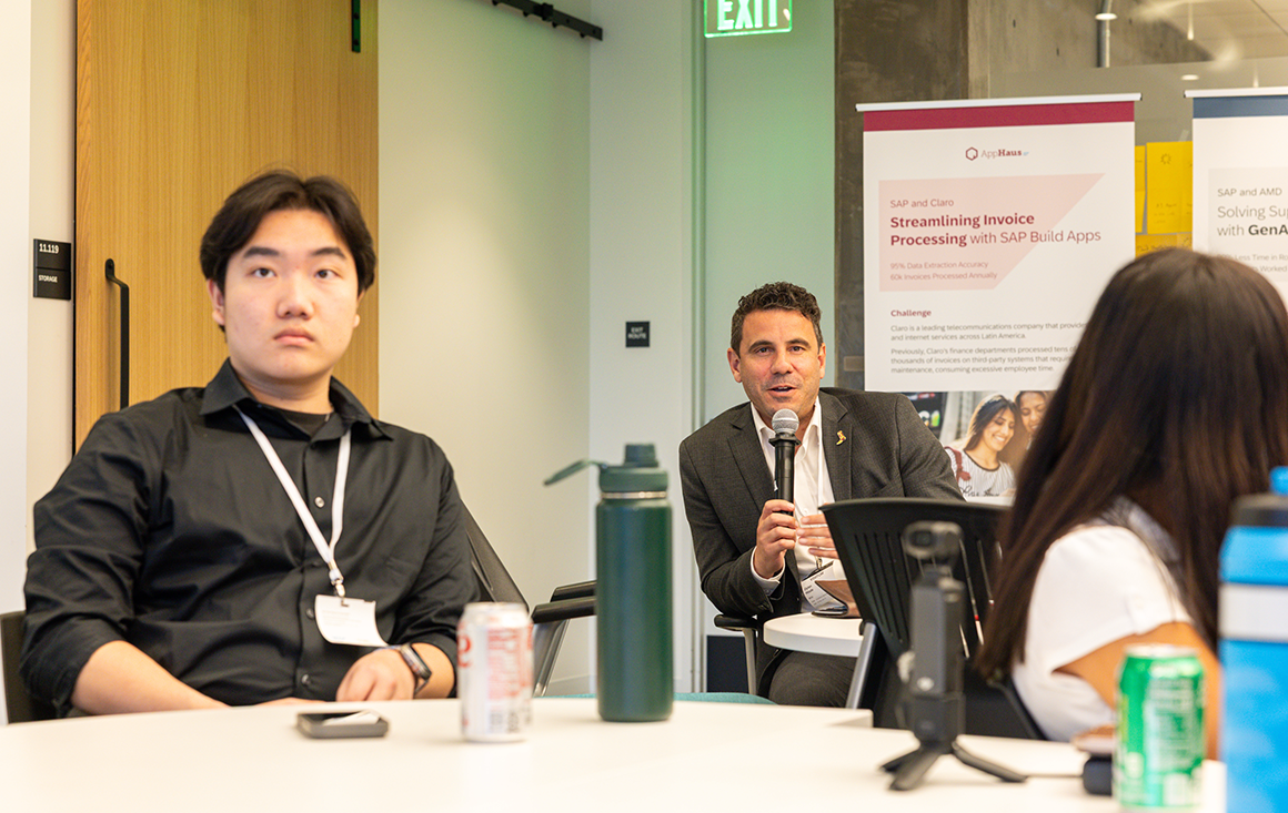 A man in a suit sits in a classroom, speaking through a mic, with students listening in the foreground.