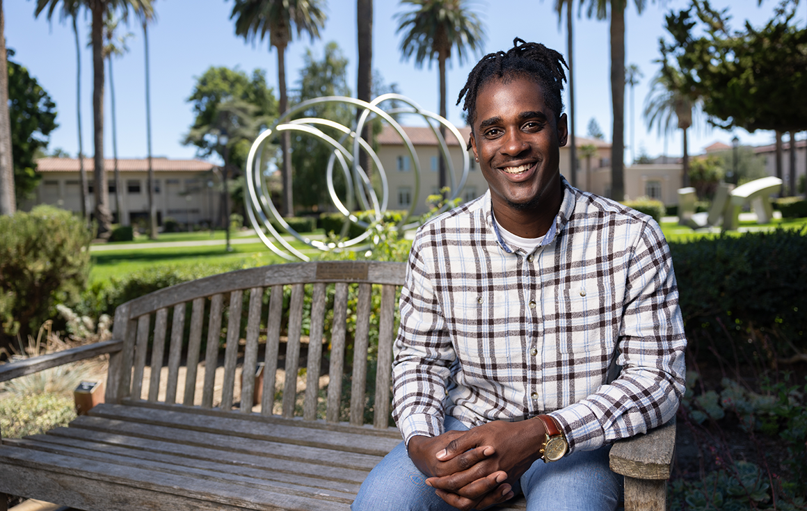 A man sits on a bench with a metal art installation of interlocking circles and palm trees in the background.