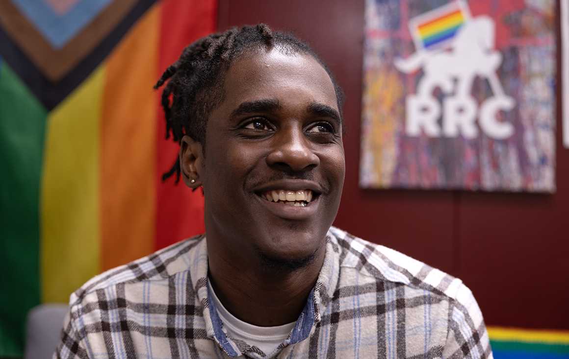 A close-up of a smiling man in a red room with rainbow flags and RRC posters on the wall behind him.