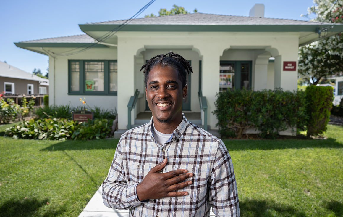 A man stands in front of a green house, holding a hand across his chest in a gesture of welcome.