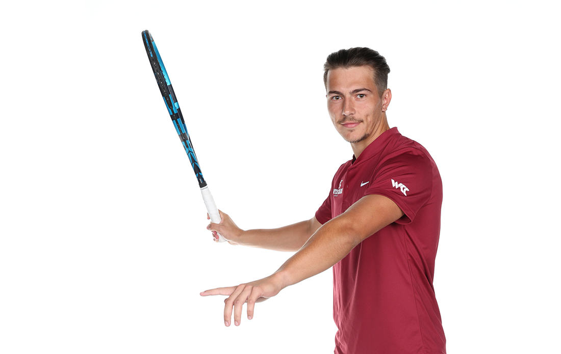 Nick Cmager holds tennis racket up at 90 degree angle while pointing with other hand.