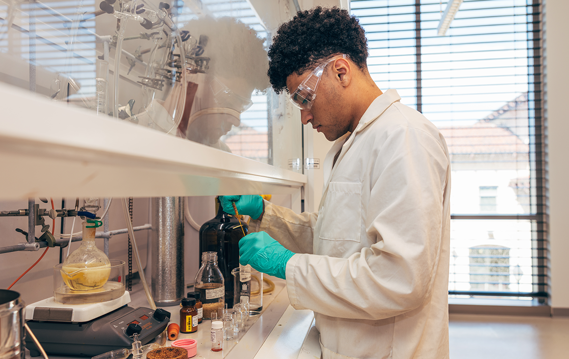 A young male student pipets solvents in a hooded lab station full of bottles and flasks.