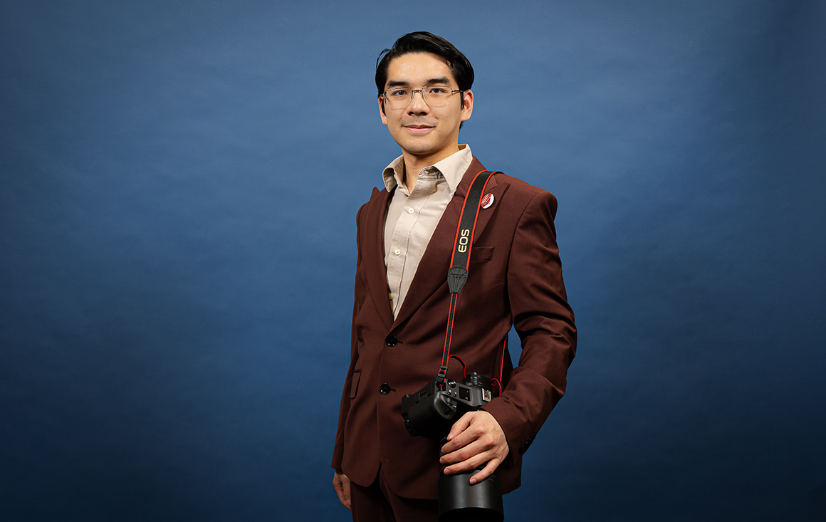 A young man in glasses and a burgundy suit holds a camera strapped over his shoulder in front of a blue backdrop.