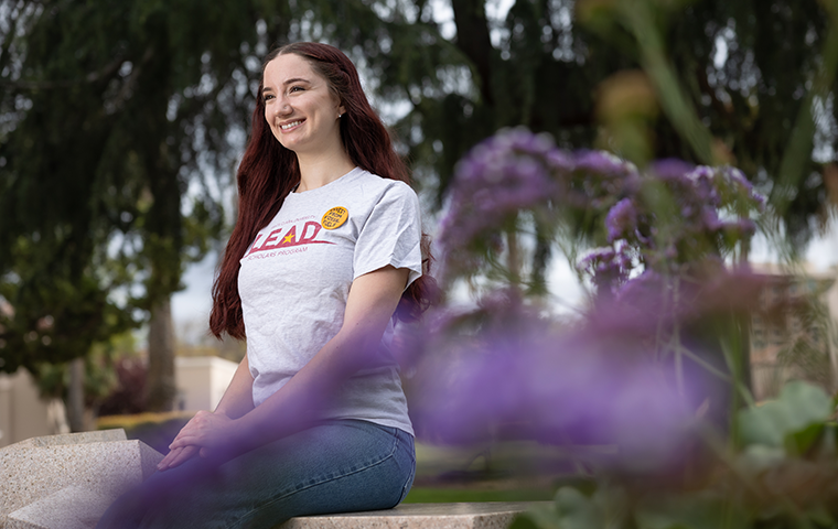 A student sits on a bench behind purple flowers in the foreground.