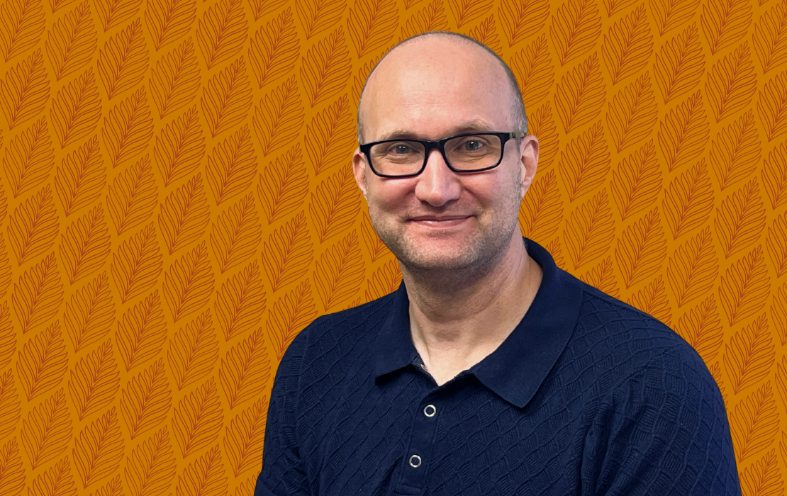 A man wearing glasses and a navy sweater posed in front of a dark gold and maroon patterned background